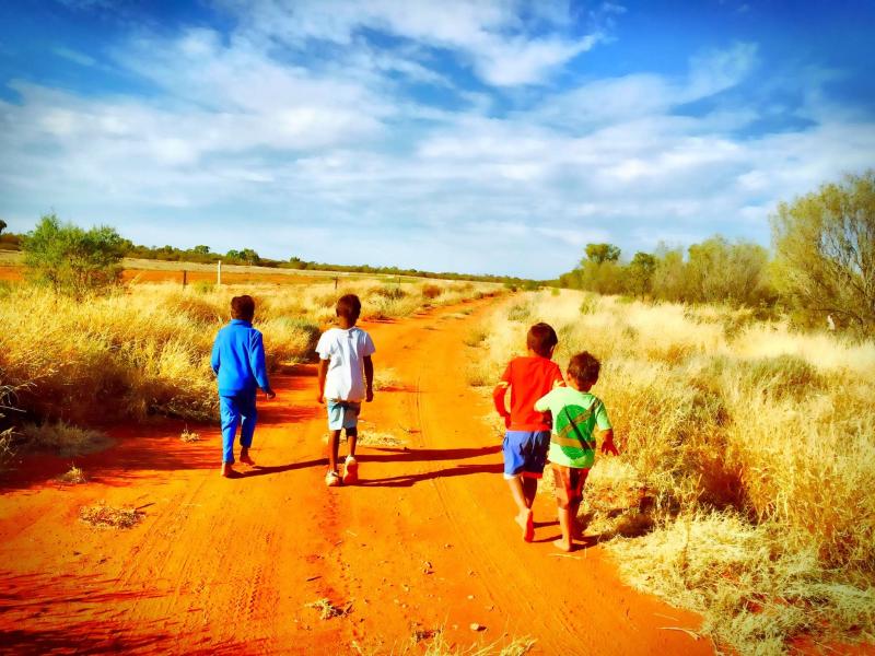 Four small children walking up a red dirt road with their backs to the camera